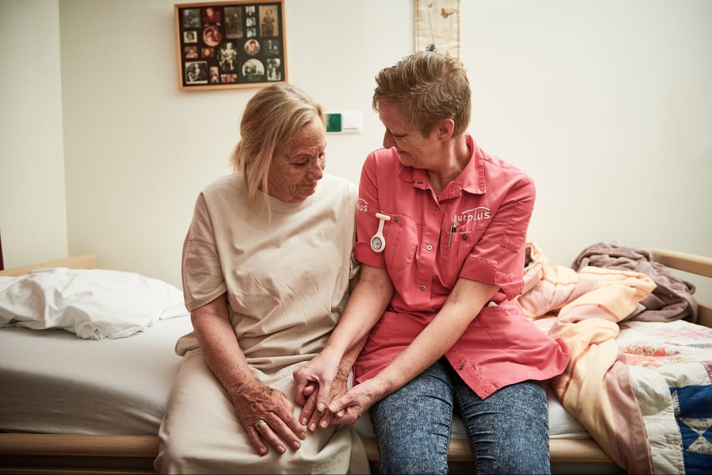 Een zorgmedewerker zit naast een bewoner op de rand van het bed